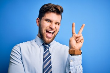 Young blond businessman with beard and blue eyes wearing elegant shirt and tie standing smiling with happy face winking at the camera doing victory sign. Number two.