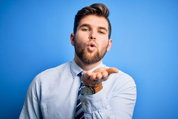 Young blond businessman with beard and blue eyes wearing elegant shirt and tie standing looking at the camera blowing a kiss with hand on air being lovely and sexy. Love expression.