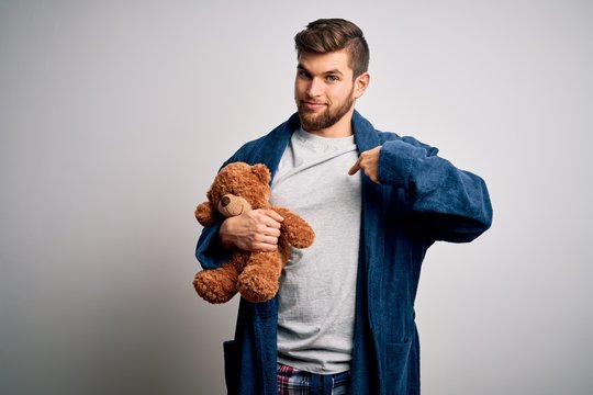 Young blond man with beard and blue eyes wearing pajama holding teddy bear with surprise face pointing finger to himself
