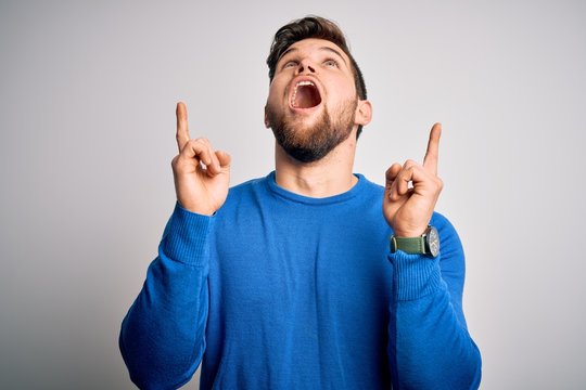 Young handsome blond man with beard and blue eyes wearing casual sweater amazed and surprised looking up and pointing with fingers and raised arms.