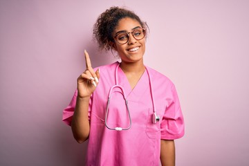 African american nurse girl wearing medical uniform and stethoscope over pink background showing and pointing up with finger number one while smiling confident and happy.