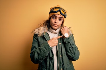 Young african american afro skier girl wearing snow sportswear and ski goggles In hurry pointing to watch time, impatience, looking at the camera with relaxed expression