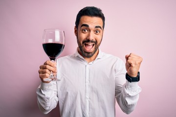 Young handsome man with beard drinking glass of red wine over isolated pink background screaming proud and celebrating victory and success very excited, cheering emotion