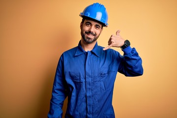 Mechanic man with beard wearing blue uniform and safety helmet over yellow background smiling doing...