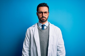 Young handsome doctor man with beard wearing coat and glasses over blue background skeptic and nervous, frowning upset because of problem. Negative person.