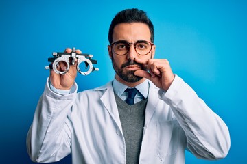 Young handsome optical man with beard holding optometry glasses over blue background mouth and lips...