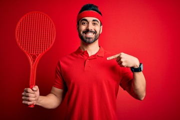 Young handsome sportsman with beard playing tennis using racket over red background with surprise face pointing finger to himself