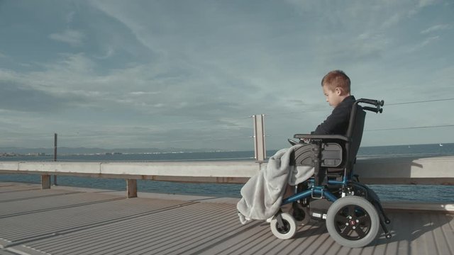 Steadicam side shot of a handicapped child in electric wheelchair riding along the waterfront