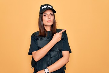 Young beautiful brunette policewoman wearing police uniform bulletproof and cap Pointing with hand finger to the side showing advertisement, serious and calm face