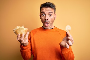 Young handsome man holding bowl with potatoes chips over isolated yellow background scared in shock with a surprise face, afraid and excited with fear expression