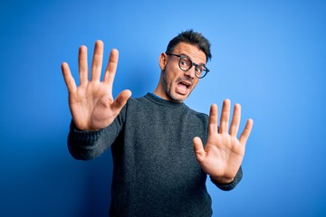 Young handsome man wearing casual sweater and glasses standing over blue background afraid and terrified with fear expression stop gesture with hands, shouting in shock. Panic concept.