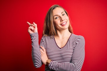 Young beautiful blonde woman wearing casual striped t-shirt over isolated red background with a big smile on face, pointing with hand and finger to the side looking at the camera.