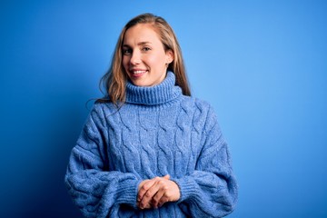 Young beautiful blonde woman wearing casual turtleneck sweater over blue background with hands together and crossed fingers smiling relaxed and cheerful. Success and optimistic