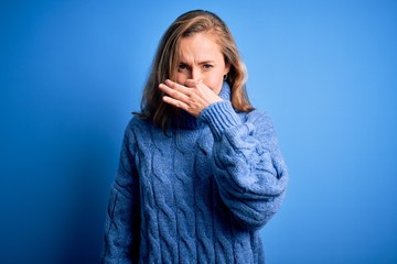 Young beautiful blonde woman wearing casual turtleneck sweater over blue background smelling something stinky and disgusting, intolerable smell, holding breath with fingers on nose. Bad smell