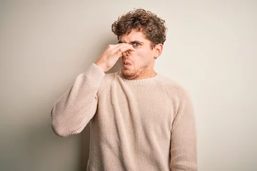 Foto op Canvas Young blond handsome man with curly hair wearing casual sweater over white background smelling something stinky and disgusting, intolerable smell, holding breath with fingers on nose. Bad smell © Krakenimages.com