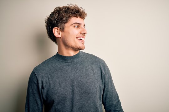 Young blond handsome man with curly hair wearing casual sweater over white background looking away to side with smile on face, natural expression. Laughing confident.