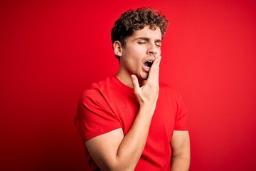Fototapeta na wymiar Young blond handsome man with curly hair wearing casual t-shirt over red background bored yawning tired covering mouth with hand. Restless and sleepiness.