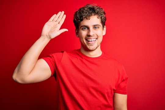 Young blond handsome man with curly hair wearing casual t-shirt over red background Waiving saying hello happy and smiling, friendly welcome gesture
