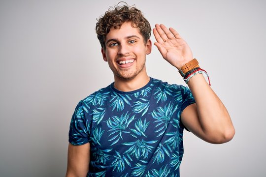 Young blond handsome man on vacation with curly hair wearing casual summer t-shirt Waiving saying hello happy and smiling, friendly welcome gesture