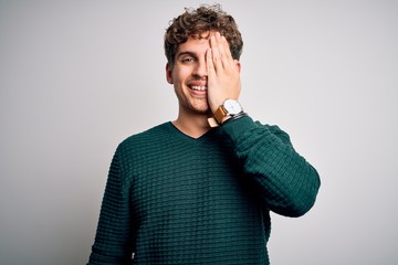 Young blond handsome man with curly hair wearing green sweater over white background covering one eye with hand, confident smile on face and surprise emotion.