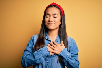 Young beautiful asian woman wearing casual denim shirt and diadem over yellow background smiling with hands on chest with closed eyes and grateful gesture on face. Health concept.