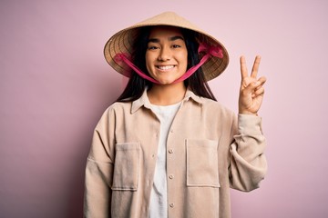 Young beautiful woman wearing traditional conical asian hat over isolated pink background smiling with happy face winking at the camera doing victory sign. Number two.