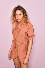 Young beautiful blonde woman with curly hair wearing casual dress standing over isolated pink background