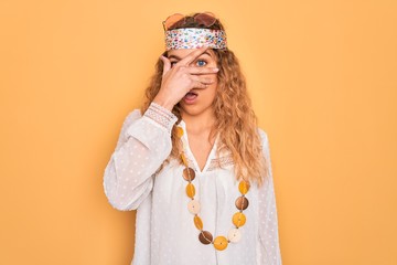 Young beautiful blonde hippie woman with blue eyes wearing sunglasses and accessories peeking in shock covering face and eyes with hand, looking through fingers with embarrassed expression.