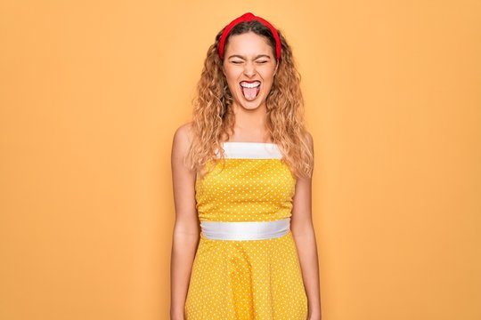Beautiful blonde pin-up woman with blue eyes wearing diadem standing over yellow background sticking tongue out happy with funny expression. Emotion concept.