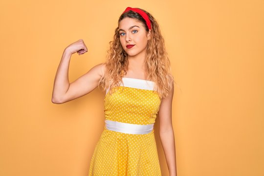 Beautiful blonde pin-up woman with blue eyes wearing diadem standing over yellow background Strong person showing arm muscle, confident and proud of power