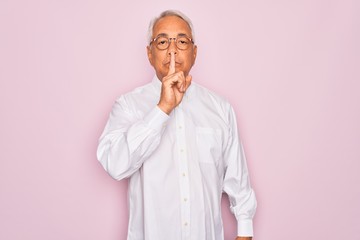 Middle age senior grey-haired man wearing glasses and business shirt over pink background asking to be quiet with finger on lips. Silence and secret concept.