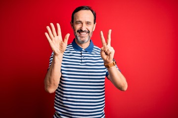 Middle age hoary man wearing casual striped polo standing over isolated red background showing and pointing up with fingers number seven while smiling confident and happy.