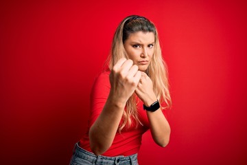 Young beautiful blonde woman wearing casual t-shirt standing over isolated red background Ready to fight with fist defense gesture, angry and upset face, afraid of problem