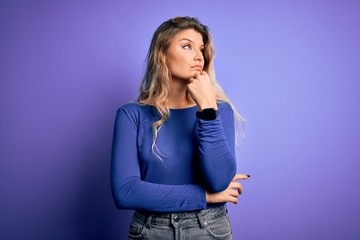 Young beautiful blonde woman wearing casual t-shirt over isolated purple background with hand on chin thinking about question, pensive expression. Smiling with thoughtful face. Doubt concept.