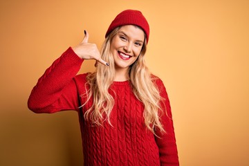 Young beautiful blonde woman wearing casual sweater and wool cap over white background smiling doing phone gesture with hand and fingers like talking on the telephone. Communicating concepts.