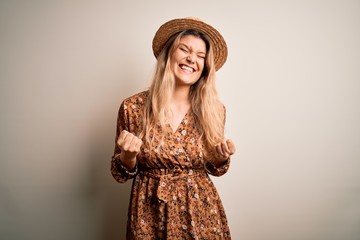 Young beautiful blonde woman wearing summer dress and hat over isolated white background very happy and excited doing winner gesture with arms raised, smiling and screaming for success. Celebration 