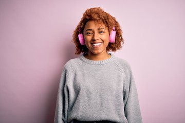 Young African American afro woman with curly hair listening to music using pink headphones with a happy and cool smile on face. Lucky person.