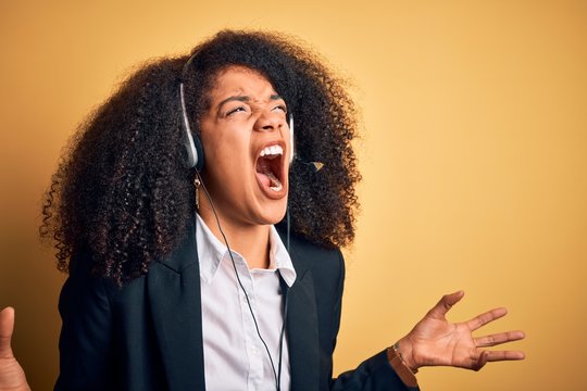 Young african american operator woman with afro hair wearing headset over yellow background crazy and mad shouting and yelling with aggressive expression and arms raised. Frustration concept.