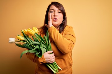 Beautiful plus size woman holding romantic bouquet of natural tulips flowers over yellow background hand on mouth telling secret rumor, whispering malicious talk conversation