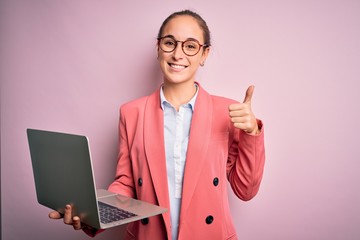 Young beautiful businesswoman wearing glasses working using laptop over pink background happy with...