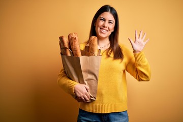Young beautiful woman holding a bag of fresh healthy bread over yellow background Waiving saying hello happy and smiling, friendly welcome gesture