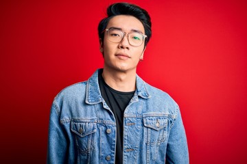 Young handsome chinese man wearing denim jacket and glasses over red background Relaxed with serious expression on face. Simple and natural looking at the camera.