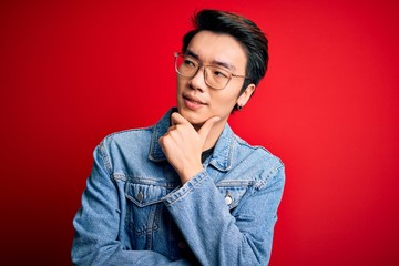 Young handsome chinese man wearing denim jacket and glasses over red background with hand on chin...