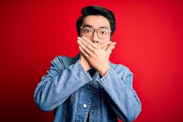Young handsome chinese man wearing denim jacket and glasses over red background shocked covering mouth with hands for mistake. Secret concept.