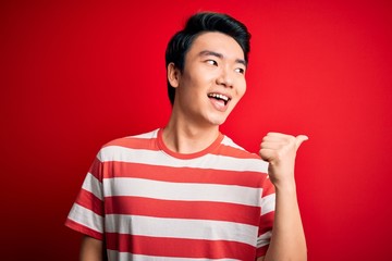 Young handsome chinese man wearing casual striped t-shirt standing over red background smiling with happy face looking and pointing to the side with thumb up.