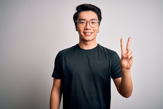 Young handsome chinese man wearing black t-shirt and glasses over white background showing and pointing up with fingers number two while smiling confident and happy.