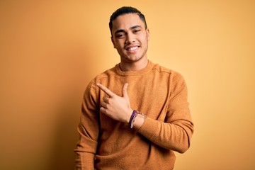 Young brazilian man wearing casual sweater standing over isolated yellow background cheerful with a smile on face pointing with hand and finger up to the side with happy and natural expression