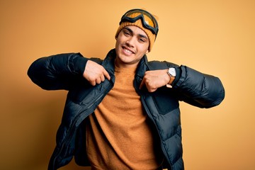 Young brazilian skier man wearing snow sportswear and ski goggles over yellow background looking confident with smile on face, pointing oneself with fingers proud and happy.