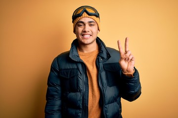 Young brazilian skier man wearing snow sportswear and ski goggles over yellow background showing and pointing up with fingers number two while smiling confident and happy.