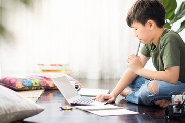 Smart looking Asian preteen boy sit crossed legs, holding pen against his lips, thinking and looking at computer laptop at home due to Covid-19 pandemic and social distancing measures